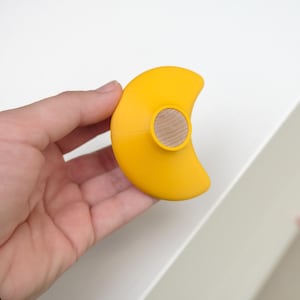 Könnte beinhalten: Gelber, halbmondförmiger Schrankknauf mit rundem Holzeinsatz. Der Knauf wird in einer Hand gehalten. Das Design ist modern und minimalistisch, vor weißem Hintergrund.