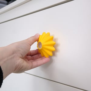 Pode incluir: Um puxador de armário amarelo brilhante em forma de flor com um centro de madeira. O puxador é segurado contra uma porta de armário branca. O design do puxador apresenta uma série de pétalas curvas, criando um efeito de raio de sol.