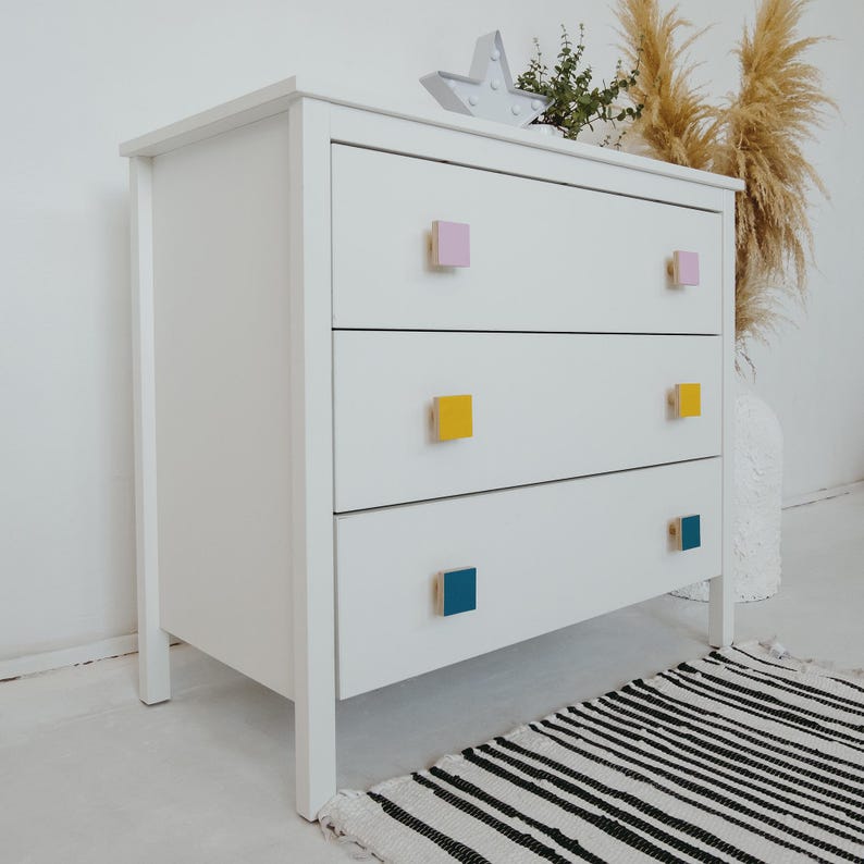 May include: White chest of drawers with four drawers and colourful square knobs. The knobs are pink, yellow and blue.