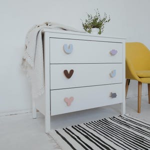 Könnte beinhalten: Eine weiße Kommode mit drei Schubladen und farbenfrohen, einzigartig geformten Schubladengriffen. Die Griffe haben die Form eines Herzens, einer Wolke, eines Vogels und anderer Formen. Eine weiße Decke ist darüber drapiert, und eine kleine Pflanze steht oben.