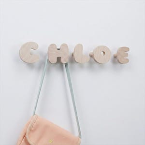 May include: A set of five light-colored wooden wall hooks shaped like the letters C, H, L, O, and E, spelling the name CHLOE. Each letter functions as an individual hook, with a pale pink fabric bag hanging from the letter H hook.