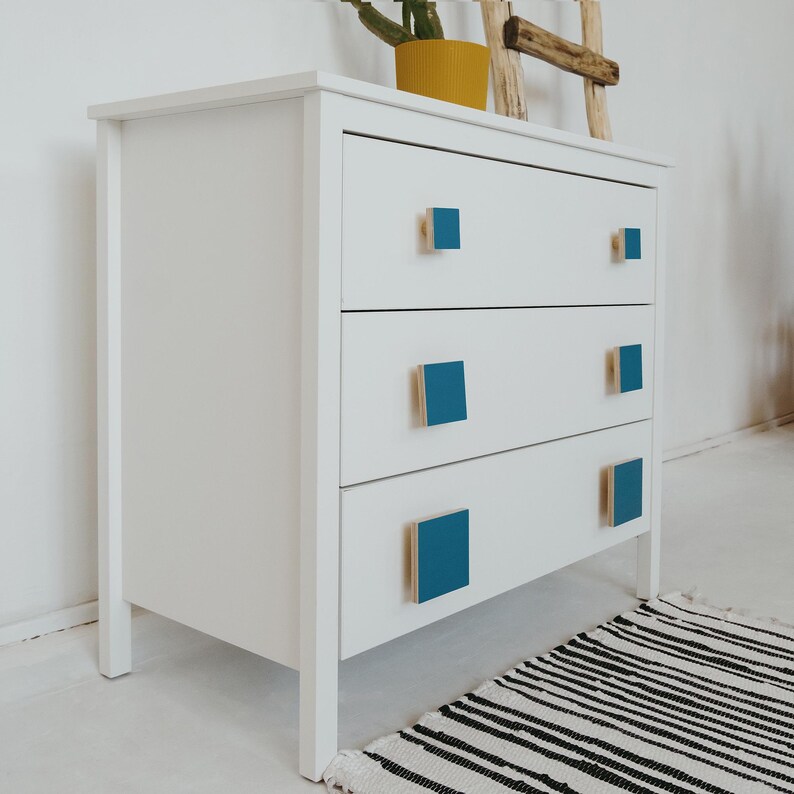 May include: White chest of drawers with three drawers and teal blue square drawer pulls. The chest of drawers is on a black and white striped rug.