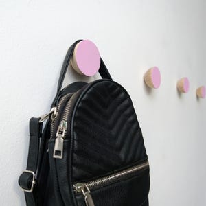 Peut inclure: Un sac à dos noir matelassé suspendu à un crochet en bois rose. Il y a trois autres crochets en bois rose sur le mur.
