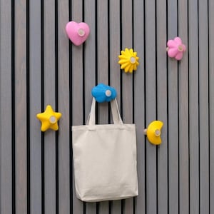 Puede incluir: Una colección de coloridos ganchos de pared con forma de estrella, corazón, flor, nube y media luna. Una bolsa de lona cuelga del gancho de nube azul. Los ganchos están montados en una pared gris con líneas verticales.