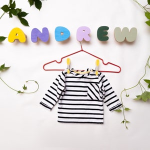 Puede incluir: Una camisa de manga larga a rayas blancas y negras con un bolsillo cuelga de una percha roja. La palabra "ANDREW" está escrita en letras de madera de colores sobre la camisa.