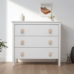 Puede incluir: Una cómoda blanca de tres cajones con pomos de madera clara. La cómoda tiene un diseño sencillo y moderno. Los pomos son redondos y de madera natural. La cómoda está colocada contra una pared blanca. Medidas en centímetros.