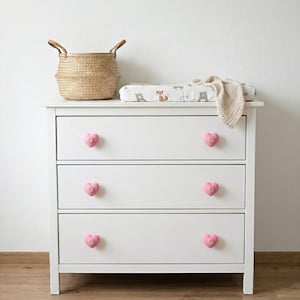 Pode incluir: Uma cômoda branca de três gavetas com puxadores em forma de coração rosa. Uma cesta de vime com alças de couro está no canto superior esquerdo, ao lado de um trocador com estampa de animais da floresta. Uma manta bege está sobre ele.