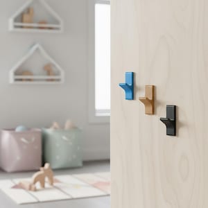 May include: Three wall hooks in blue, tan, and black are mounted on a light wood surface. The hooks have a simple, modern design. The background shows a room with shelves, storage bins, and a rug.