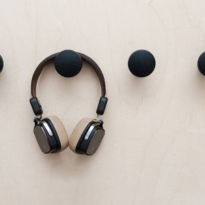 Peut inclure: Quatre patères murales en bois noir sont fixées sur une surface en bois clair. Un casque audio beige et noir est suspendu à l'un des crochets. Les crochets sont espacés uniformément.