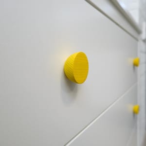 Puede incluir: Primer plano de una cómoda blanca con tres cajones, cada uno con un pomo de madera amarillo brillante en forma de semicírculo. Los pomos están espaciados uniformemente, creando un contraste. La veta de la madera es visible.