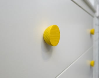Puede incluir: Primer plano de una cómoda blanca con tres cajones, cada uno con un pomo de madera amarillo brillante en forma de semicírculo. Los pomos están espaciados uniformemente, creando un contraste. La veta de la madera es visible.