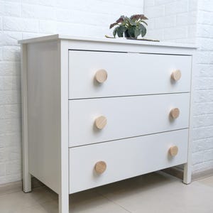 May include: A white three-drawer dresser with light wood knobs. The dresser has a simple, modern design and is set against a white brick wall. A small potted plant sits on top.