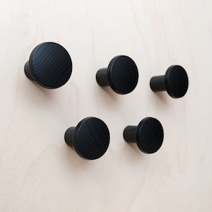 May include: Five black wooden wall hooks with a rounded, slightly textured surface. The hooks are mounted on a light wood background.