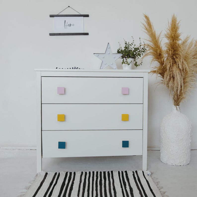 May include: White chest of drawers with three drawers and colourful square drawer pulls. The top drawer has pink pulls, the middle drawer has yellow pulls, and the bottom drawer has teal pulls.