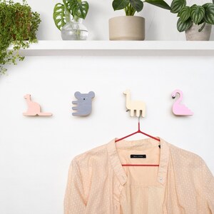 May include: Four wooden animal-shaped hooks in pink, grey, and beige, mounted on a white wall. The hooks are shaped like a kangaroo, koala, llama, and flamingo. A red hanger with a beige shirt with white polka dots hangs from the llama hook.