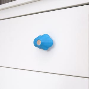 Könnte beinhalten: Blauer wolkenförmiger Schrankknauf mit hellem Holzmittelstück, an einer weißen Schublade befestigt. Der Knauf ist ein dekoratives Element, das Möbeln einen skurrilen Touch verleiht. Die Schublade ist Teil eines weißen Schranks.