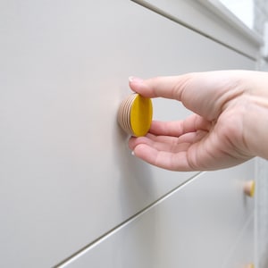 Puede incluir: Un pomo de cajón de madera y amarillo con un patrón rayado. El pomo está siendo presionado por una mano.
