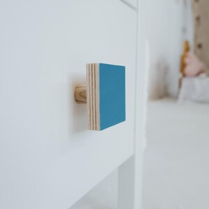 May include: A square cabinet knob with a light blue front and a natural wood back. The knob is mounted on a white surface.