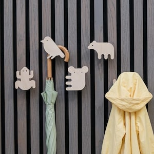 Puede incluir: Cuatro ganchos de pared de madera clara con forma de pájaro, rana, oso y koala. Un paraguas verde claro cuelga del gancho de pájaro y una chaqueta amarilla con capucha cuelga del gancho de oso, montados sobre una pared de listones verticales.