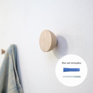 Pode incluir: Um gancho de parede de madeira clara com um design arredondado de meio círculo. O gancho está montado numa parede branca. A imagem também mostra uma bucha de parede azul e um parafuso, com o texto "the set includes".