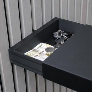 Peut inclure: Une étagère murale rectangulaire noire avec un tiroir coulissant. À l'intérieur du tiroir ouvert, on trouve un billet de 100 hryvnias, plusieurs pièces de monnaie argentées et un porte-clés en métal, le tout fixé sur un mur à lattes grises.