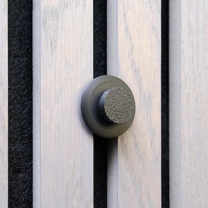 May include: A close-up of a dark gray, textured knob attached to a light gray wooden panel. The panel has vertical, evenly spaced wooden slats. The background is a dark, textured material.
