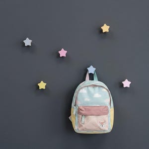Può includere: Uno zaino per bambini con un design a nuvole e volpe è appeso a un gancio a forma di stella. Ulteriori ganci a forma di stella in colori pastello sono montati su una parete grigio scuro. Lo zaino ha una combinazione di colori azzurro, rosa e giallo.
