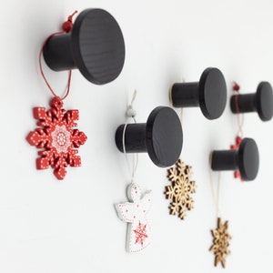 May include: Five black wooden hooks with rounded tops are mounted on a white wall. Each hook has a string hanging from it with a decorative ornament attached. The ornaments include a red snowflake, a white angel, and three wooden snowflakes.