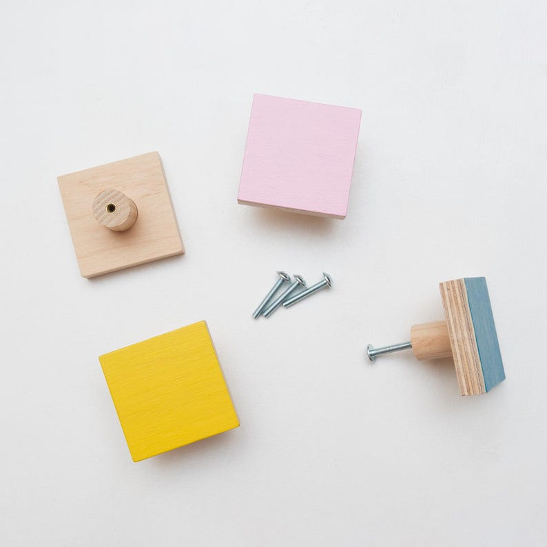 May include: Four wooden knobs with different coloured paint finishes: natural wood, yellow, pink and blue. The knobs are square shaped and have a small hole in the centre for mounting.