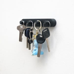 Puede incluir: Una taza de cerámica blanca con asa negra y el texto "But First, Coffee" en negrita negra. La taza se encuentra sobre un fondo borroso, sugiriendo un ambiente acogedor.