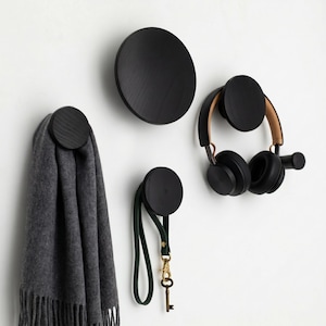 May include: A set of four circular black wooden wall hooks mounted on a wall. One hook holds a grey fringed scarf, another holds a dark green leather strap with a metal key, and a third hook supports a pair of black over-ear headphones.
