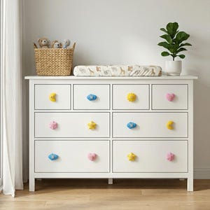 Könnte beinhalten: Eine weiße Kommode mit bunten, geformten Knöpfen, darunter gelbe Monde, blaue Wolken, gelbe Sterne und rosa Herzen. Eine Wickelauflage mit Tiermuster liegt obenauf, zusammen mit einem Weidenkorb mit Stofftieren und einer Topfpflanze.