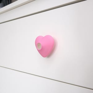 Pode incluir: Um puxador de armário rosa em forma de coração com uma inserção de madeira, montado em uma porta de armário branca. O puxador é de cor rosa lisa e lisa, e a inserção de madeira é visível no centro do coração.