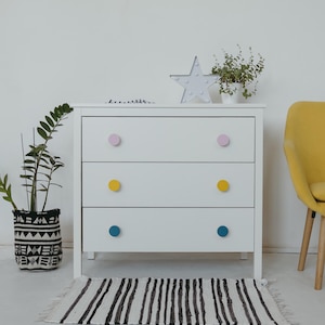 Puede incluir: Una cómoda blanca de tres cajones con pomos de colores. Los pomos son rosas, amarillos y azules. Una alfombra a rayas blancas y negras está frente a la cómoda. Una luz en forma de estrella y plantas en macetas están encima.