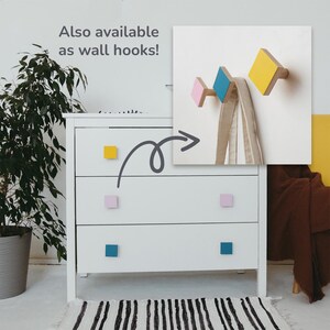 May include: A white chest of drawers with three drawers, each with a different coloured square knob. The knobs are yellow, pink, and blue. The dresser is in front of a white wall with a picture of three wooden wall hooks painted in pink, blue, and yellow. The text "Also available as wall hooks!" is written on the wall above the picture.