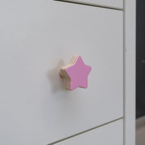 May include: A pink star-shaped cabinet knob with a natural wood base is attached to a white cabinet. The knob is painted pink on the front and sides, with the natural wood grain visible on the edges. The knob is a decorative accent.