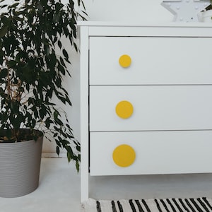 May include: A white three-drawer dresser with bright yellow circular knobs. The dresser is set against a white wall, with a potted plant to the left. A black and white striped rug is visible at the bottom.