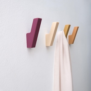 Peut inclure: Ensemble de quatre patères murales de couleurs différentes : prune, crème, moutarde et rouille. Un foulard clair est accroché à la patère crème. Les patères sont fixées sur un mur blanc. Elles sont en bois.