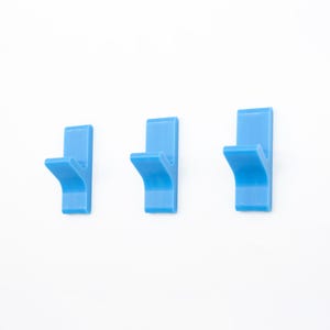 May include: Three light blue wall hooks are shown against a white backdrop. Each hook features a rectangular base and a curved hook. The hooks are evenly spaced, providing a simple, modern design for organizing items in a room.