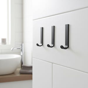Peut inclure: Trois crochets noirs en métal en forme de J fixés sur un mur blanc. Les crochets sont espacés uniformément et ont un design minimaliste. L'arrière-plan montre une salle de bain avec un lavabo.