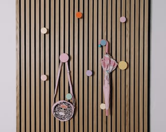 Può includere: Una parete a doghe di legno con ganci rotondi colorati. Una borsetta rosa con un disegno di bambola è appesa a un gancio rosa. Anche un ombrello rosa e bianco con un design a cartoni animati è appeso.