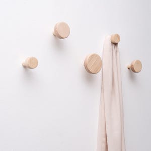 Może przedstawiać: Cztery jasne, drewniane wieszaki ścienne o okrągłym kształcie zamontowane na białej ścianie. Jeden wieszak trzyma jasnoróżowy szalik. Wieszaki są równomiernie rozmieszczone, oferując minimalistyczny i funkcjonalny design.