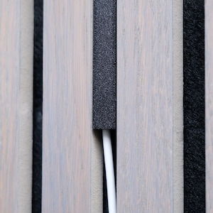 Può includere: Primo piano di un design di pannelli in legno con doghe verticali in legno grigio chiaro. Una striscia nera strutturata si trova tra le doghe di legno. Un cavo bianco corre verticalmente attraverso un'apertura nel pannello.