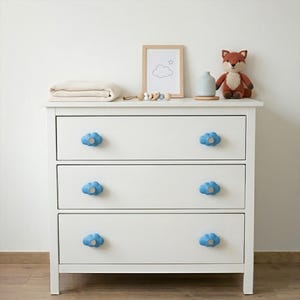 Puede incluir: Una cómoda blanca de tres cajones con pomos azules en forma de nube. La parte superior de la cómoda muestra un cuadro enmarcado, un zorro de peluche, un pequeño jarrón, una manta doblada y un juguete de madera. Mueble de guardería.