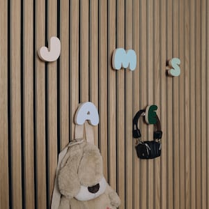 May include: A wooden wall with a set of colorful wooden letter hooks spelling out the name "JAMES". A plush toy is hanging from the "A" hook. A pair of headphones is hanging from the "E" hook.