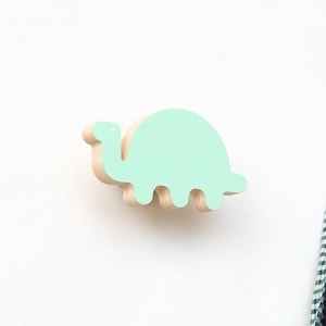 May include: A light green wooden dinosaur wall hook. The dinosaur is shaped like a stegosaurus with a long neck and a rounded body. The hook is made of plywood and has a natural wood finish.