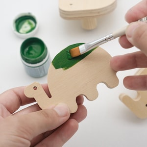 May include: A flat, light-colored wooden cutout shaped like a dinosaur being painted with a flat-tipped brush dipped in vibrant green paint. A small jar of green paint sits nearby, and another wooden dinosaur shape is visible in the background.