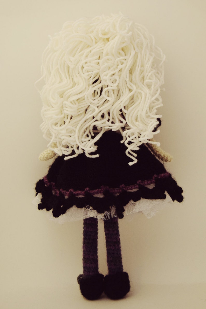 May include: A crocheted doll with long white yarn hair, a black dress with a white lace trim, purple legs, and black shoes.
