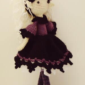 May include: Crocheted doll with white yarn hair, a black dress with purple trim, and purple yarn legs. The doll has a purple flower on its head and black eyes.