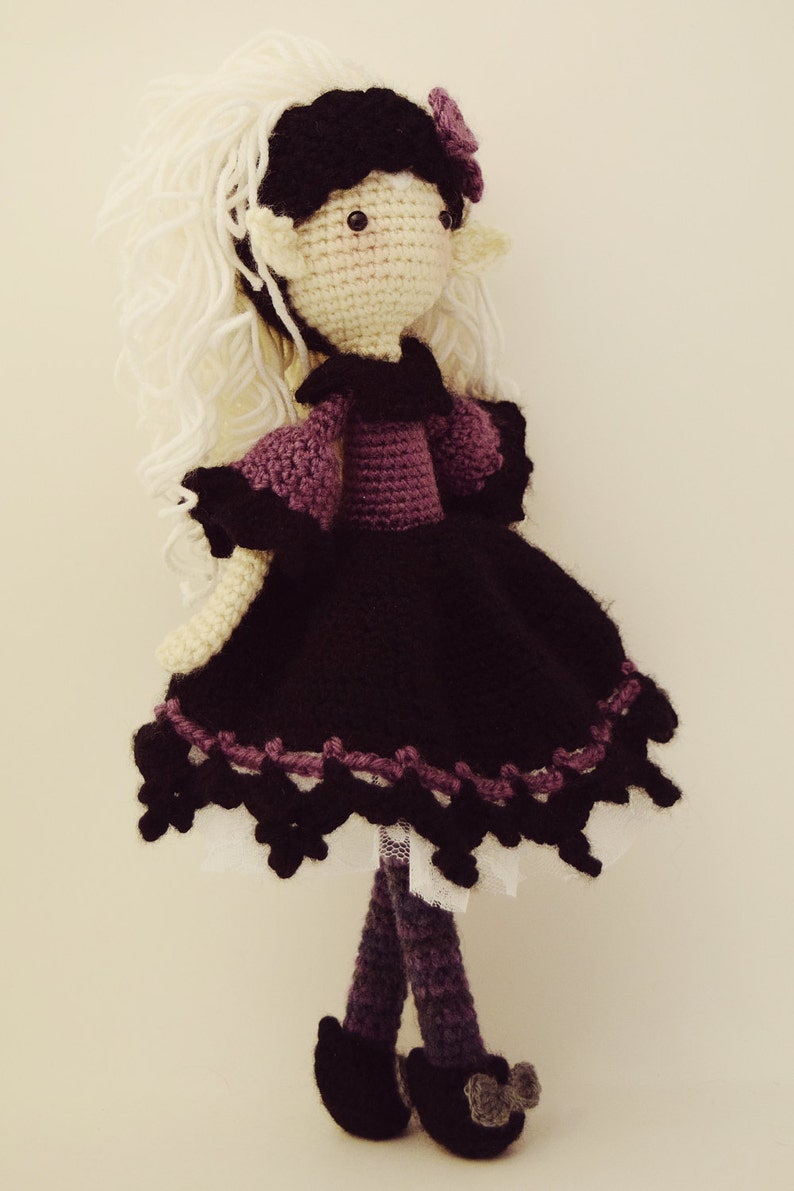 May include: Crocheted doll with white yarn hair, a black dress with purple trim, and purple yarn legs. The doll has a purple flower on its head and black eyes.